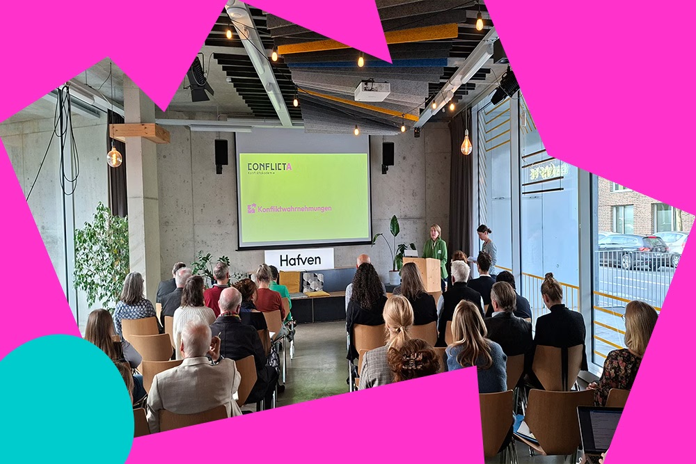 Eine Gruppe von Menschen sitzt in einem Veranstaltungsraum und schaut auf einen großen Bildschirm mit der Präsentationsfolie ‚CONFLICTA – Konfliktwahrnehmungen‘. Am Podium stehen zwei Personen, und eine davon spricht.