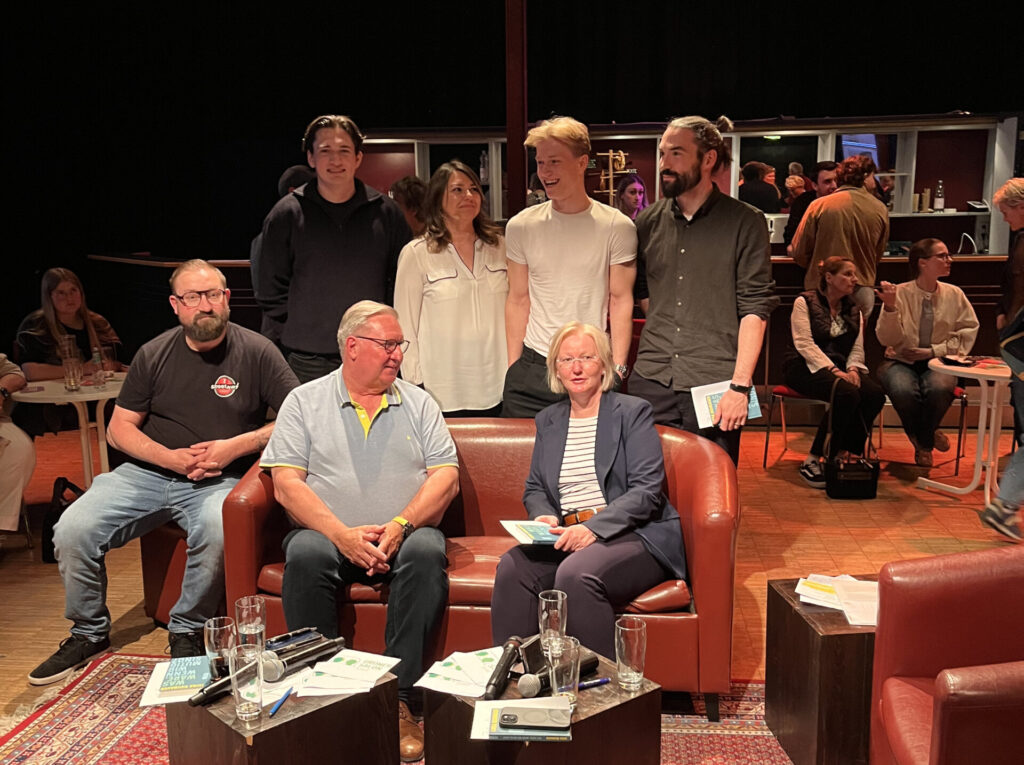 Gruppenbild der fünf Podiumsteilnehmer*innen und zwei Moderator*innen, in erster Reihe sitzend zu dritt, dahinter stehend zu viert. Im Hintergrund: Publikum, welches im Saal ankommt und Bar.
