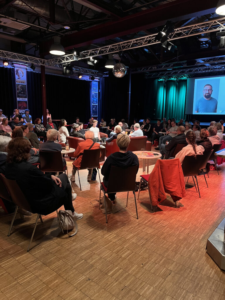 Das Podium sitzt zentral im Saal auf Sofas, das Publikum darum verteilt. Alle Blicke sind auf die laufende PowerPoint-Präsentation gerichtet, die die Podiumsteilnehmer*innen vorstellt.