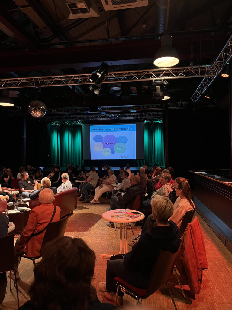 Publikum aus den hintersten Reihen blickt auf Präsentation, Folie stellt unterschiedliche Ebenen einer Definition des Begriffs „Jugenden“ vor. Folie zeigt die Kategorien „biologisch“, „politisch“, „rechtlich“, „sozial“ und „institutionell“.