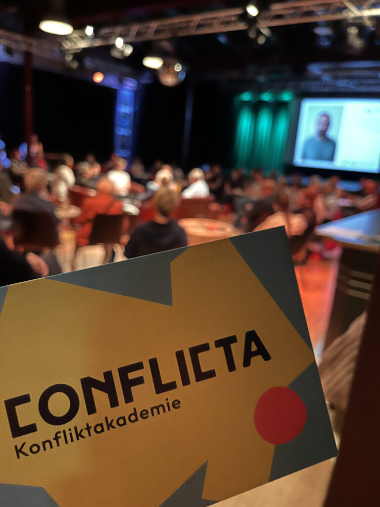 Im Vordergrund, scharf: Postkarte der ConfliktA mit offiziellem Logo. Im Hintergrund, unscharf: Der mit Publikum gefüllte Saal.