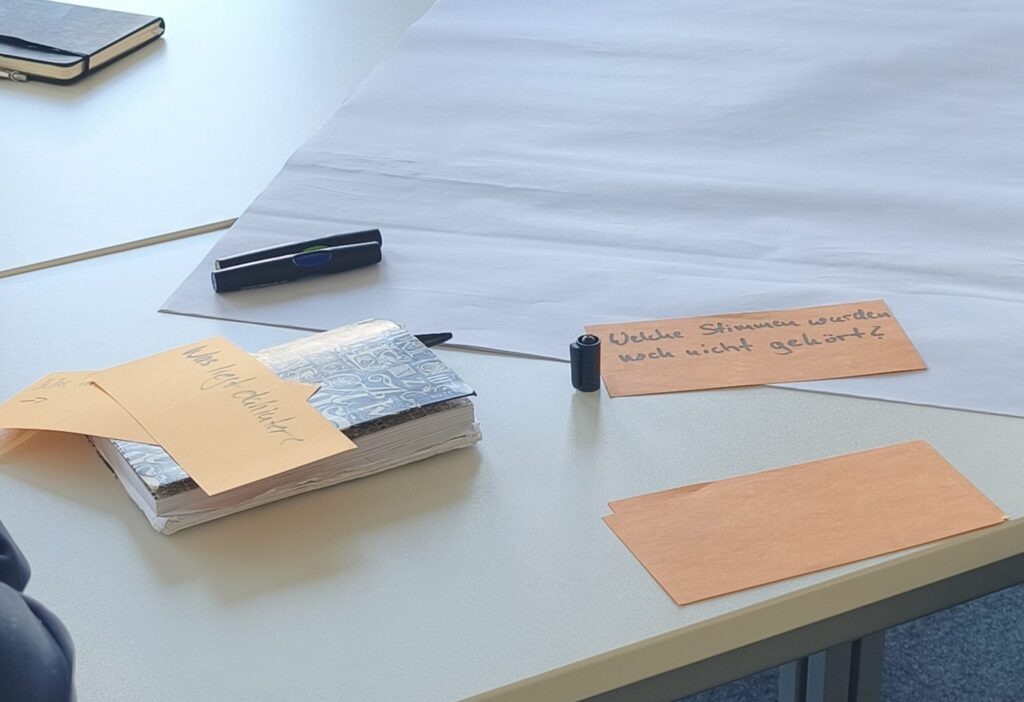 Symbolbild: Tisch mit zwei Notizbüchern, and und Arm von zwei Personen sind am Bildrand erkennbar. Orange Karten und schwarze Textmarker liegen mittig, auf einer der Karten steht "Welche Stimmen wurden noch nicht gehört?"