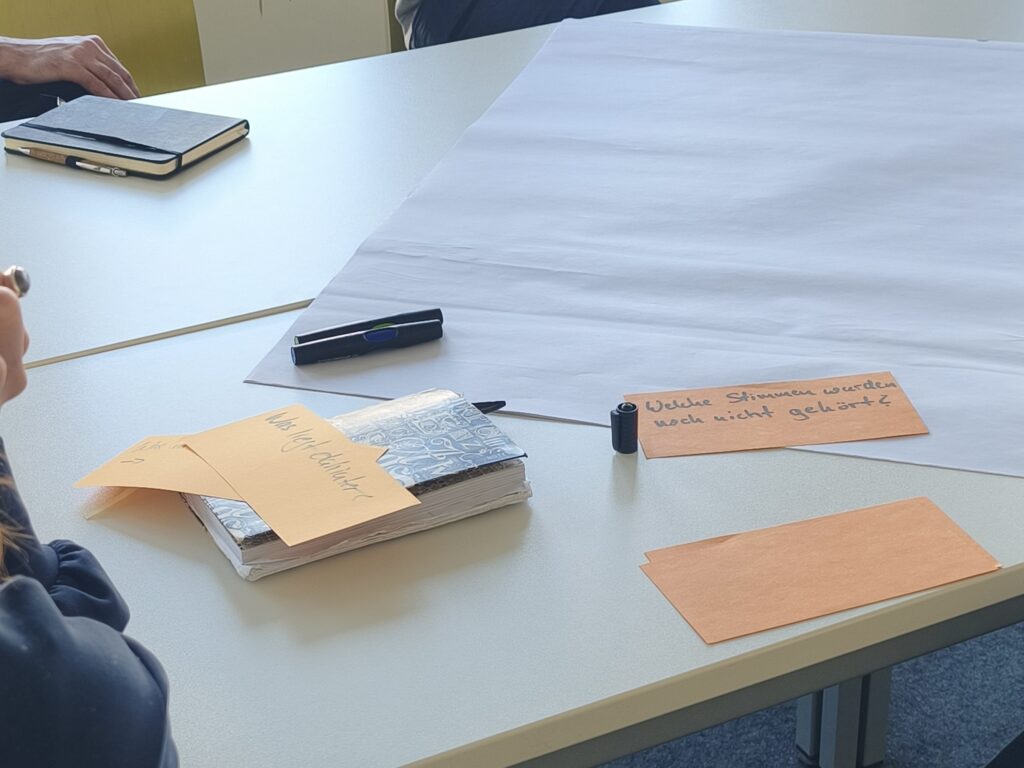 Symbolbild: Tisch mit zwei Notizbüchern, and und Arm von zwei Personen sind am Bildrand erkennbar. Orange Karten und schwarze Textmarker liegen mittig, auf einer der Karten steht "Welche Stimmen wurden noch nicht gehört?"