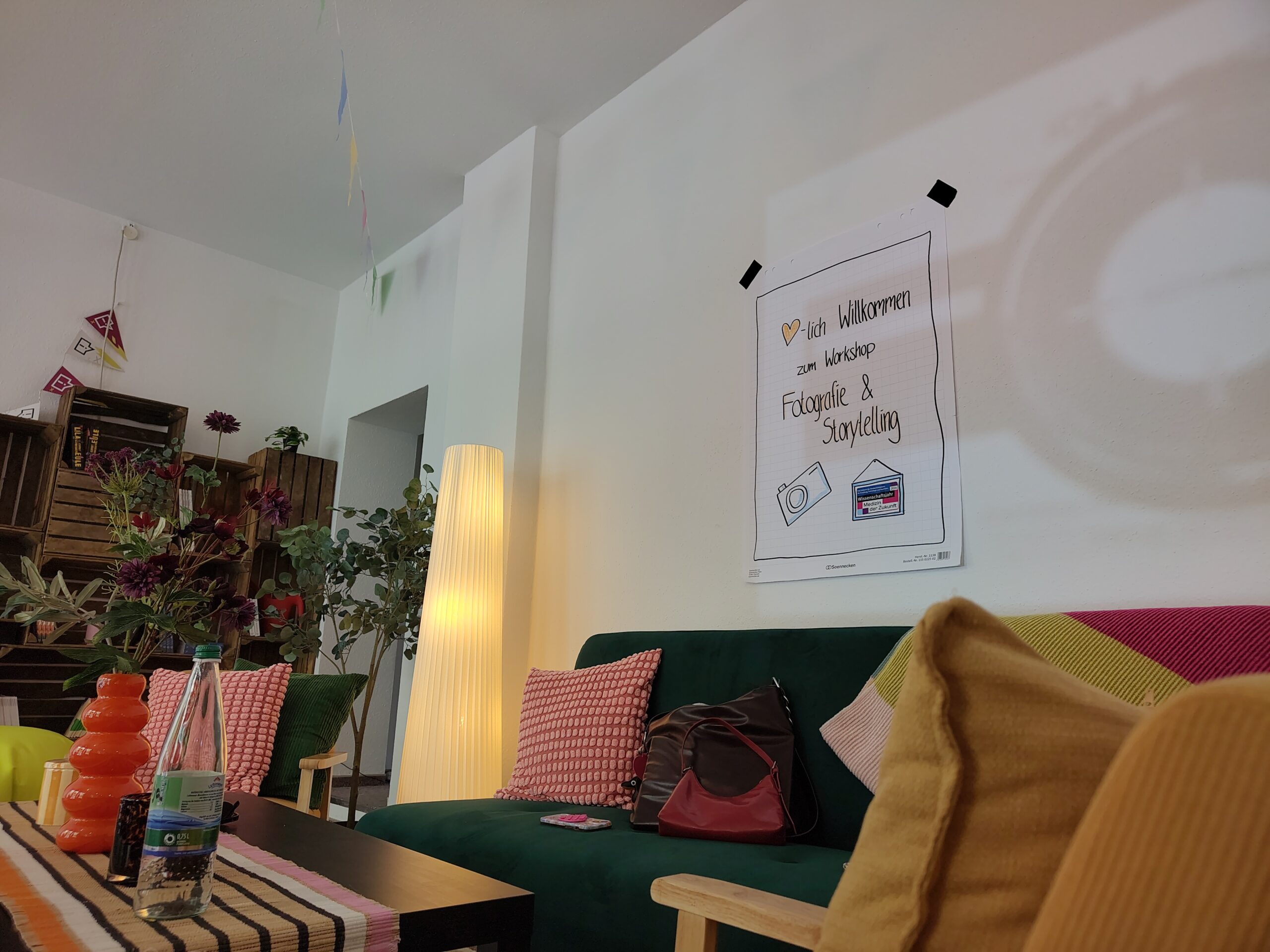 Foto von einem gemütlich und bunt eingerichteten Raum. An der Wand hängt ein Plakat mit der Aufschrift: Herzlich willkommen zum Workshop Fotografie und Storytelling.