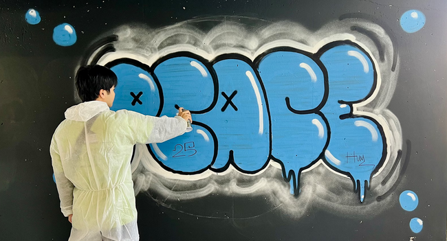 Eine Person sprüht mit einer Sprühdose ein "Peace" (Frieden) in blauer Farbe an eine Wand.