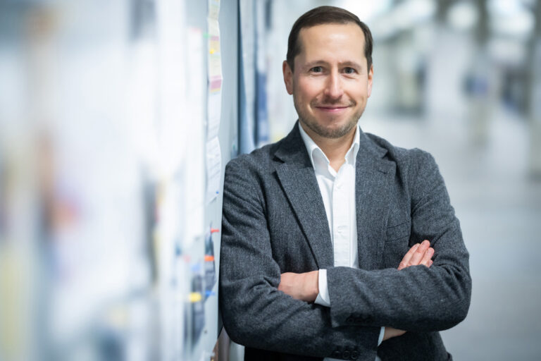 Andreas Uhl steht in einem hellen Flur angelehnt an eine Wand mit verschränkten Armen.