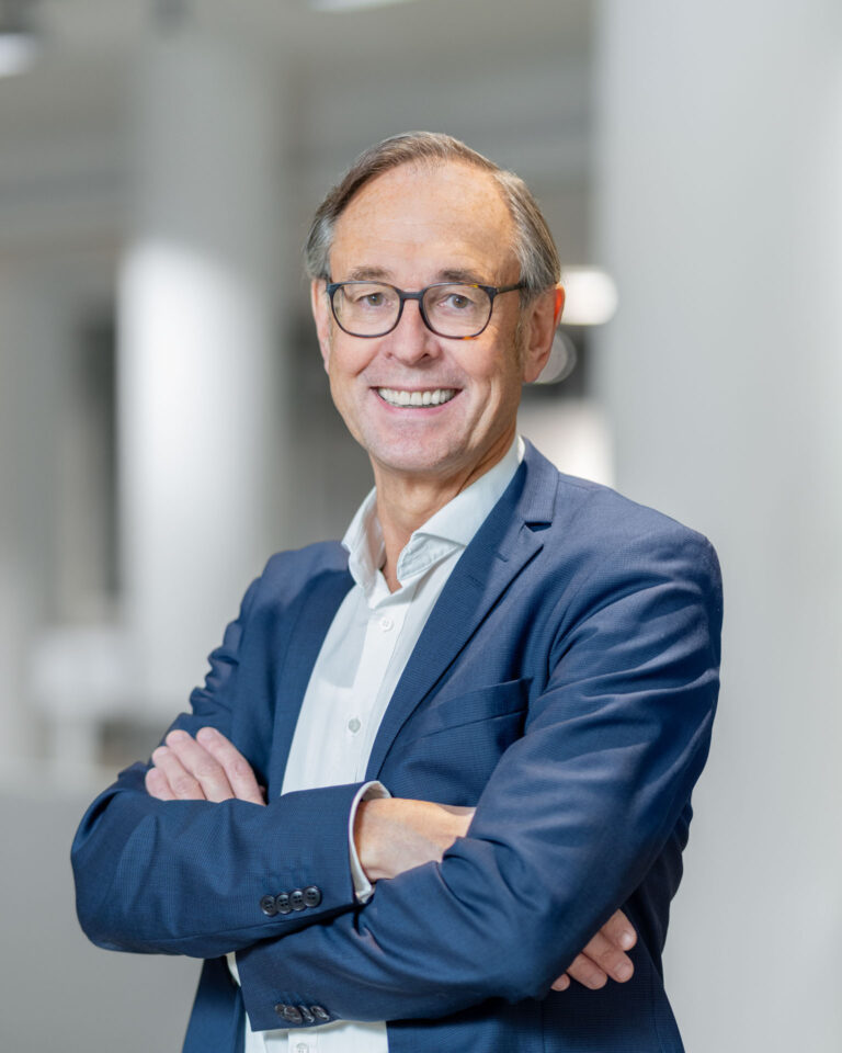 Prof. Dr. Andreas Zick steht mit verschränkten Armen in einem hellen Flur und lächelt.