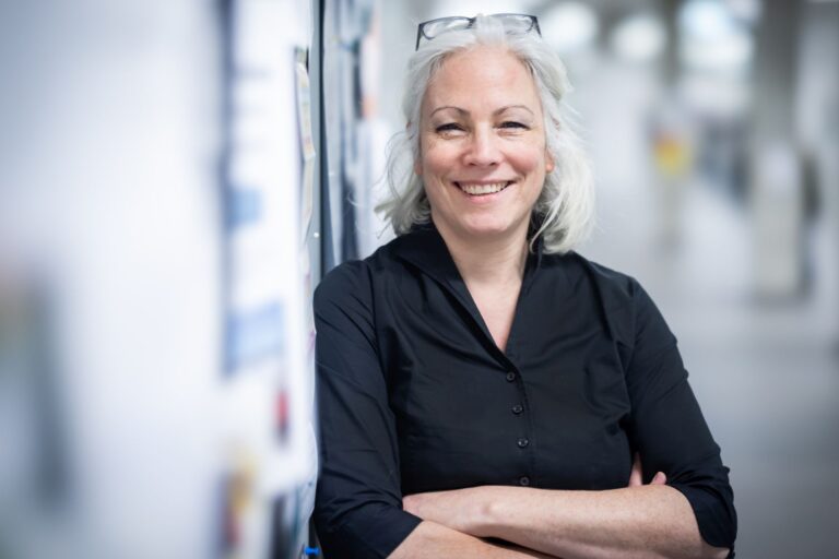 Dr. Kerstin Eppert steht mit verschränkten Armen in einem hellen Flur, lehnt an einer Wand und lächelt.